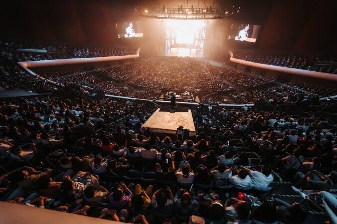 Arena llena vista desde lo alto, con el escenario iluminado y pantallas