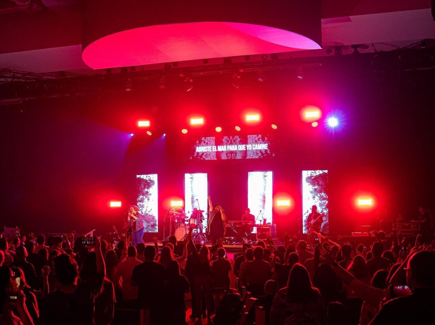 Escenario con luces rojas y pantallas, frente al público con las manos en alto