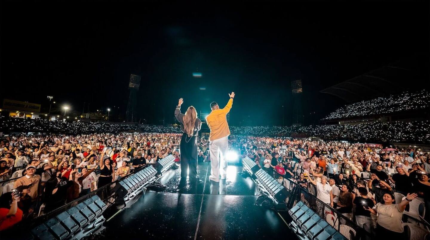 Dos artistas de espaldas en el escenario, con los brazos en alto, frente a un estadio lleno iluminado por los celulares
